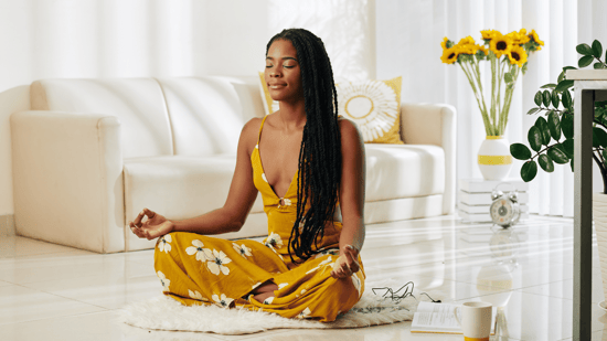 black woman with long hair in yellow jumpsuit with white flowers sitting on floor meditating in a light, airy room
