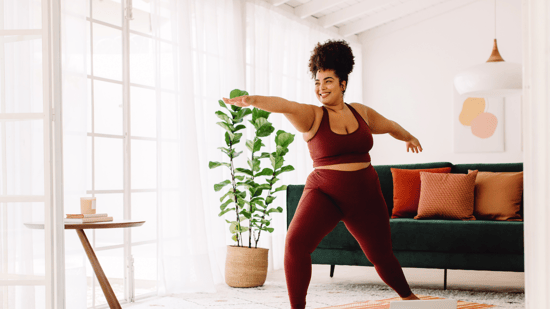 black woman wearing burgundy workout clothing standing in living room doing yoga pose